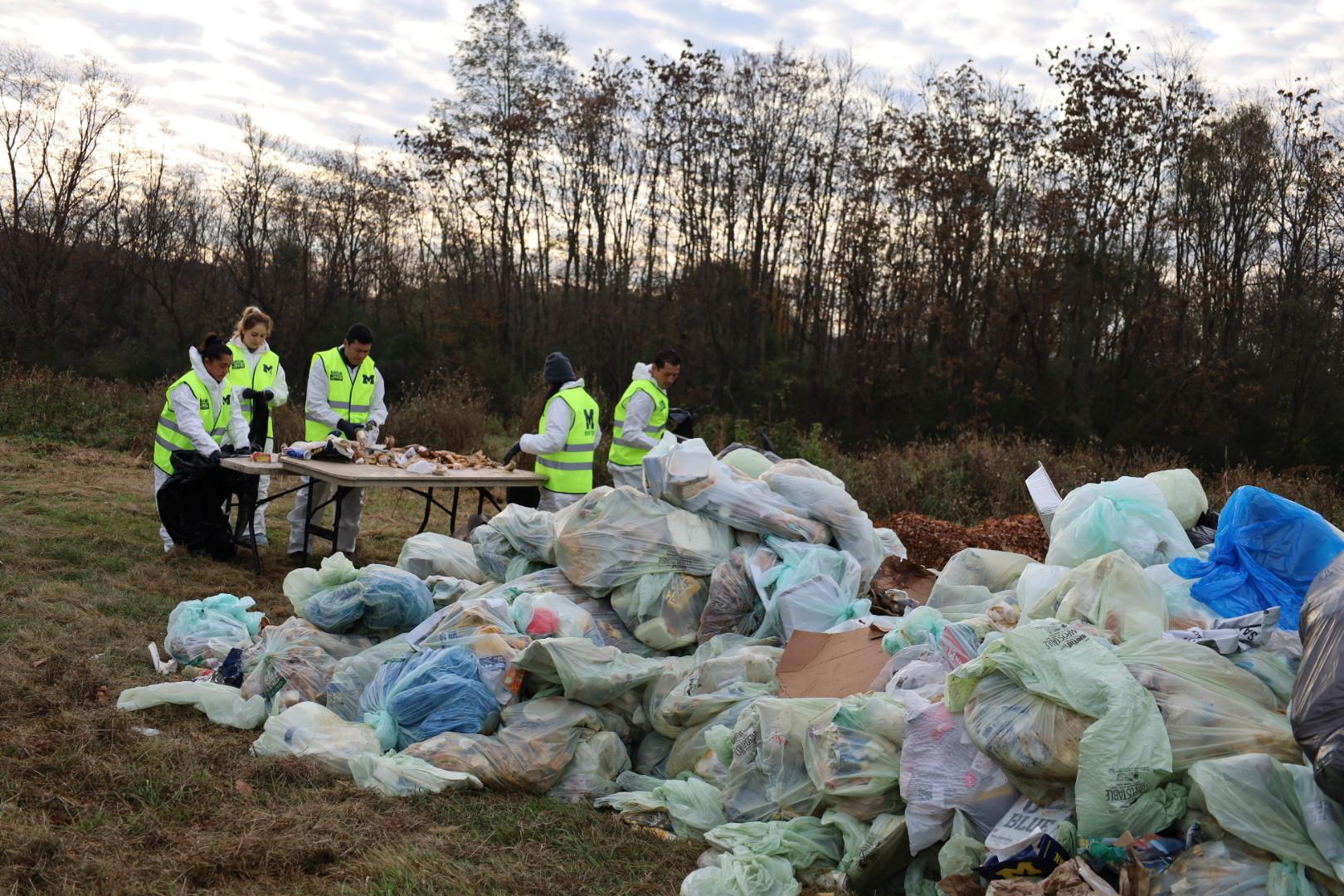 5 people stand at a table sorting compostable materials, a large pile of filledgarbage bags is behind them