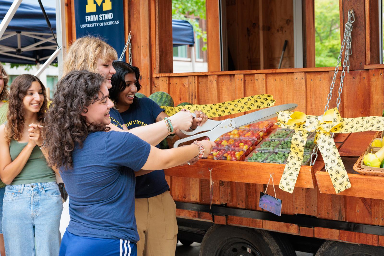 Three students hold a large pair of scissors and cut a yellow ribbon from the new Farm Stand, a fourth student clasps their hands and smiles.