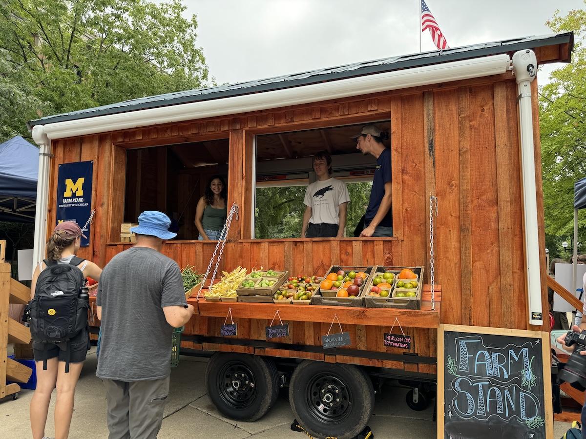 far stand with three students inside and two shoppers buying produce