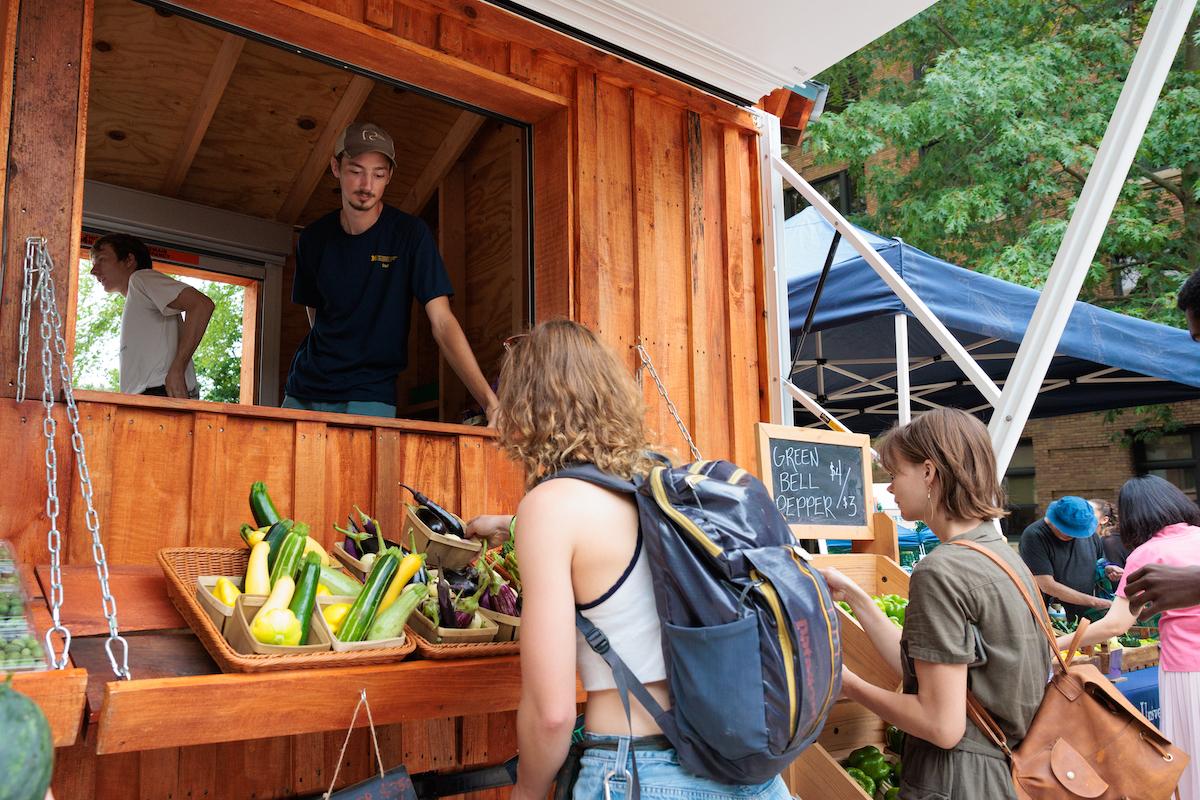 student in farm stand looking at customers browsing produce