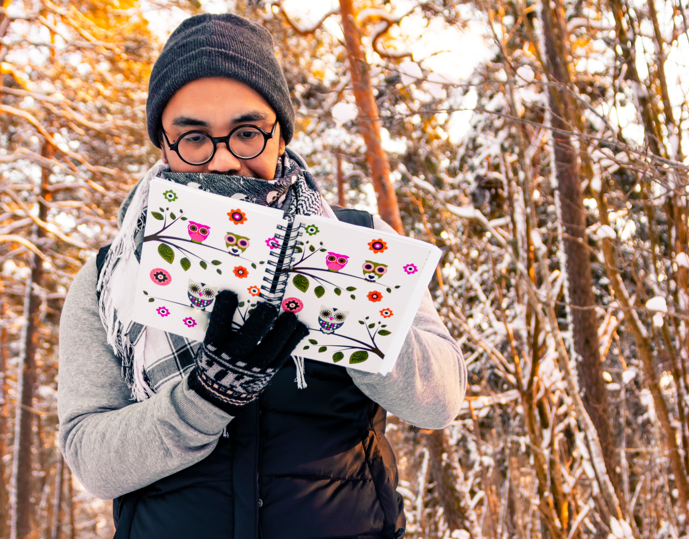 person outside in winter with trees behind them holding a journal with flowers and owls drawn on it