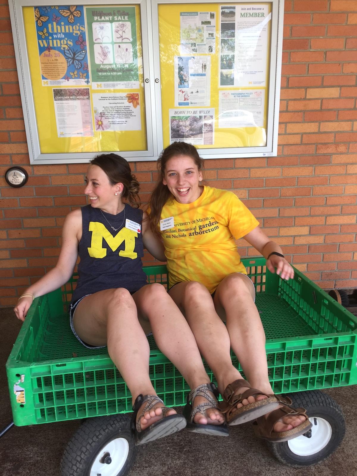 Two MBGNA interns laughing as they sit in a green yard cart