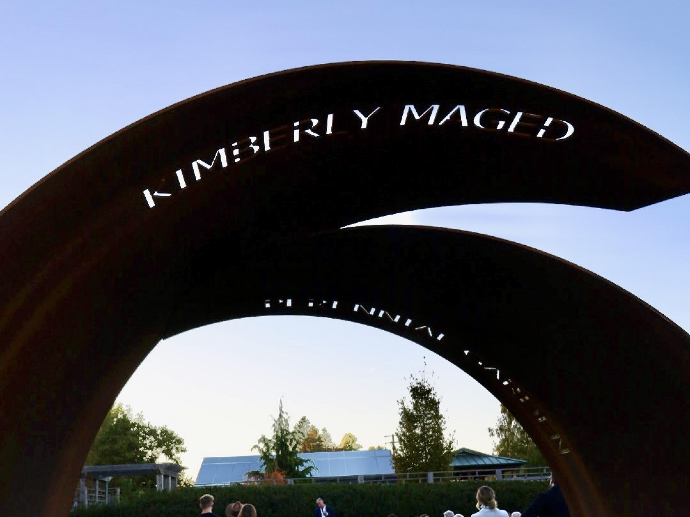 Archway into perennial garden taht says Kimberly Maged Perennial Garden.  Beyond the archway we can see a gathering with Theodore Maged at the center, giving remarks