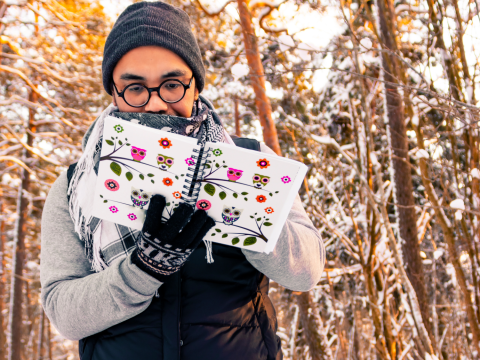 person outside in winter with trees behind them holding a journal with flowers and owls drawn on it