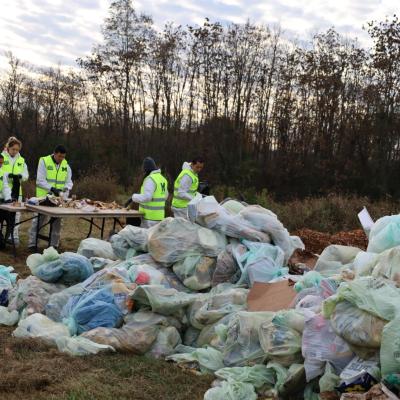 5 people stand at a table sorting compostable materials, a large pile of filledgarbage bags is behind them