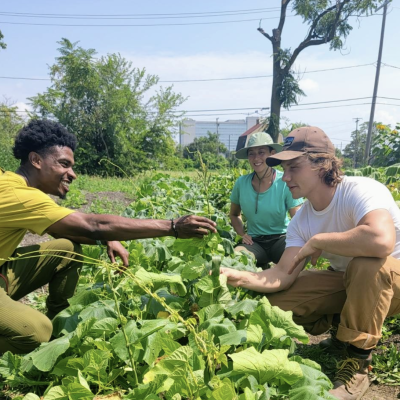 three people in a garden field and one hands another a vegetable