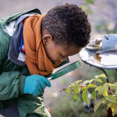 A child leans over with a hand held magnifying glass looking at a plant 